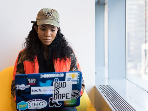 Young woman wearing a cap and camo jacket working on a laptop covered with tech and motivational stickers in a bright office.